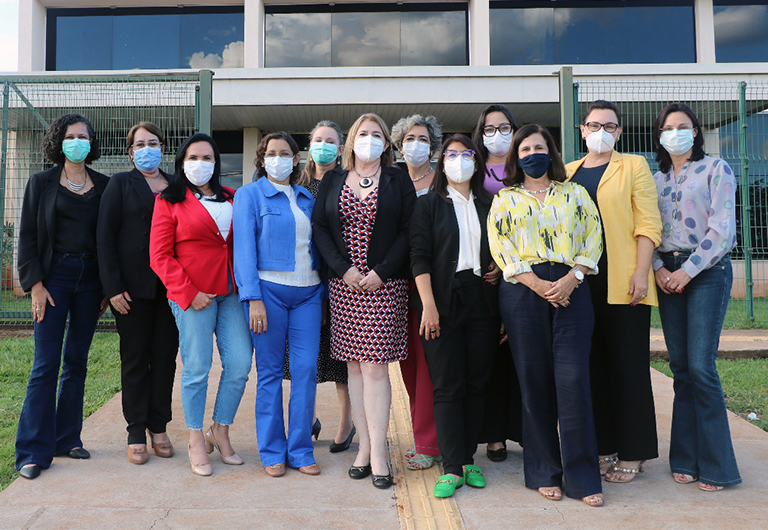 Doze mulheres aparecem na foto. Todas elas estão de máscara em frente a um prédio com janelas de vidro.